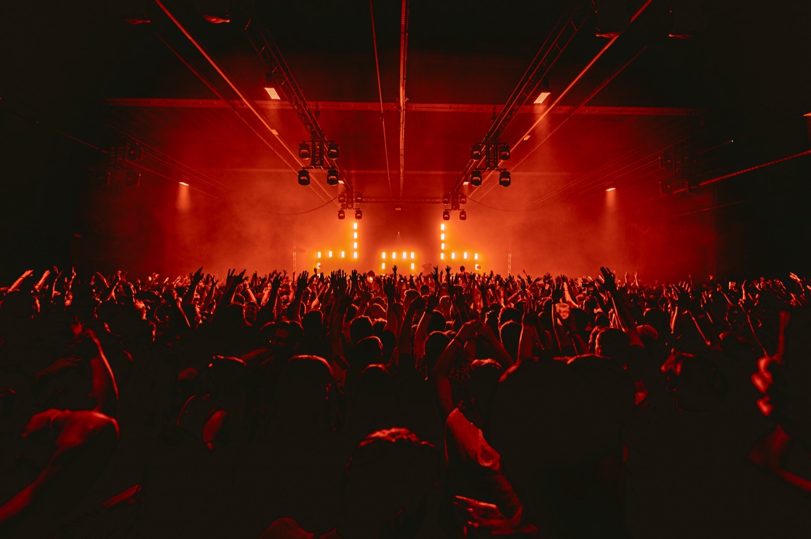 Indoor crowd under red lights at Antwerp Expo, hands in the air